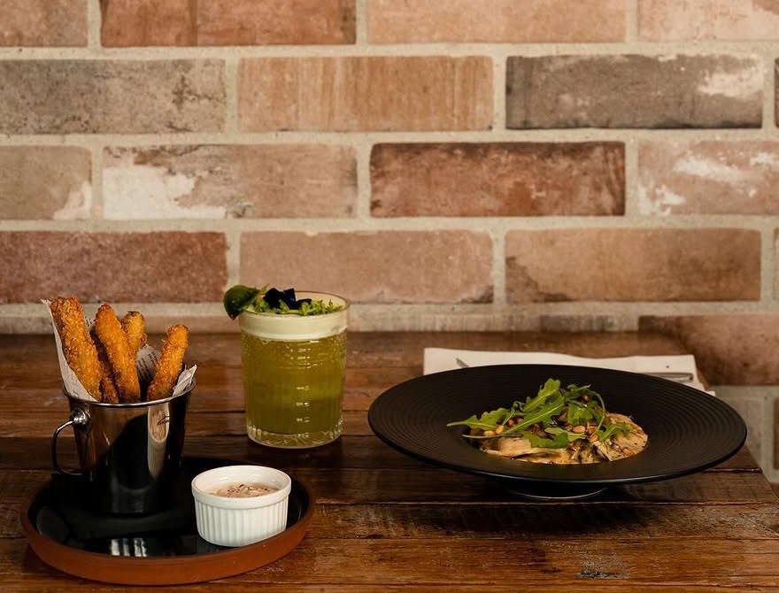 Bebida verde, petiscos panados com molho e um prato com rúcula numa mesa de madeira