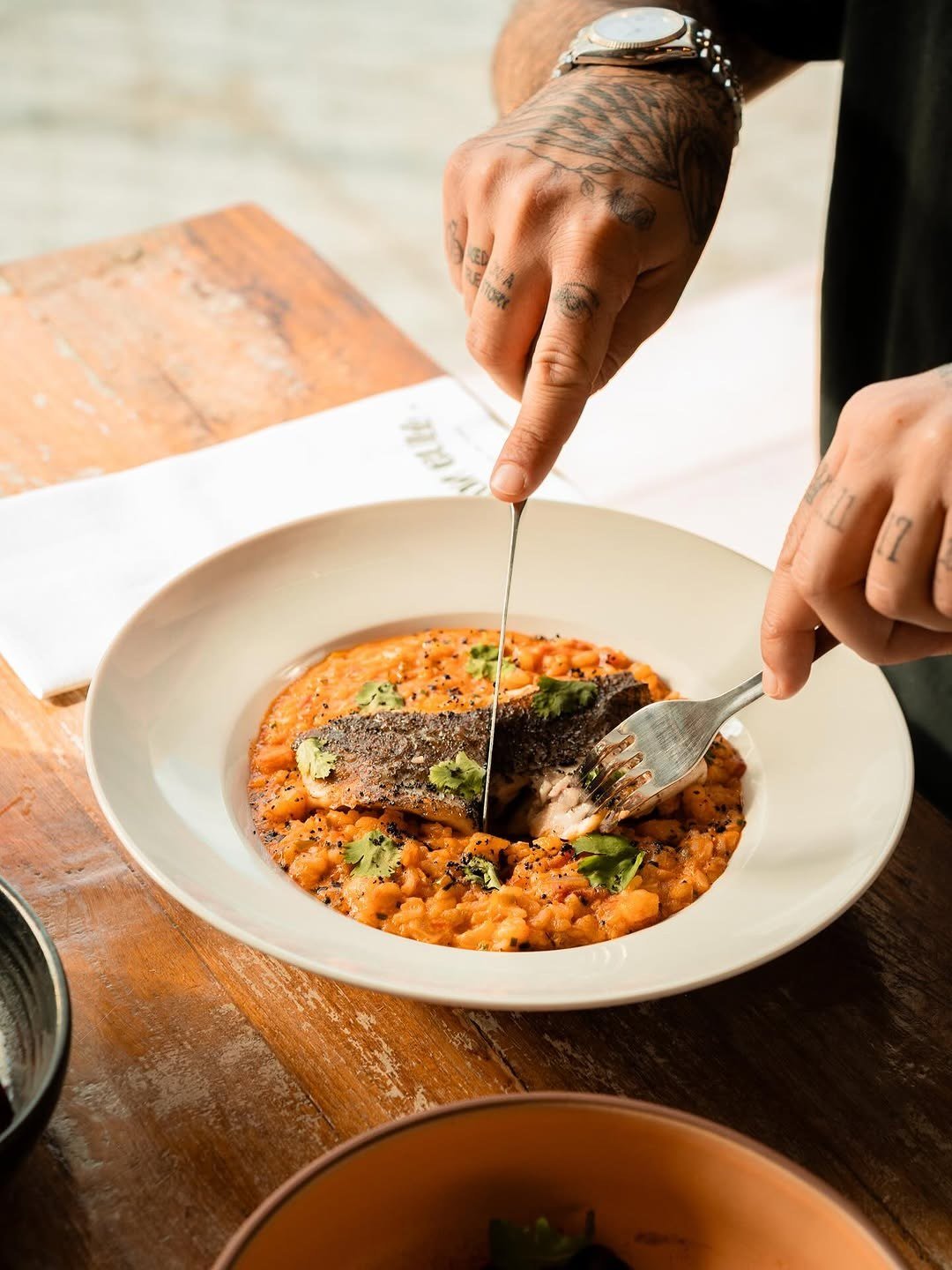 Peixe sobre arroz alaranjado num prato fundo branco, numa mesa de madeira